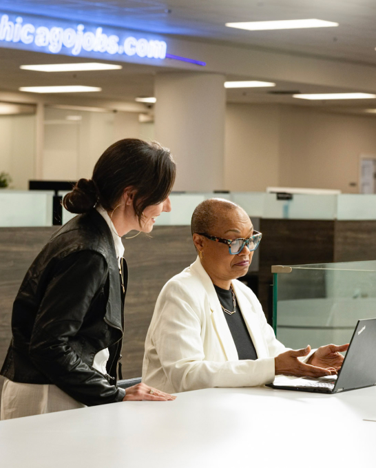 Two people are working in an office. One is seated, wearing glasses and a white blazer, typing on a laptop. The other is standing, wearing a black jacket, and looking at the laptop screen. A blue neon sign "chicagojobs.com" is visible in the background.
