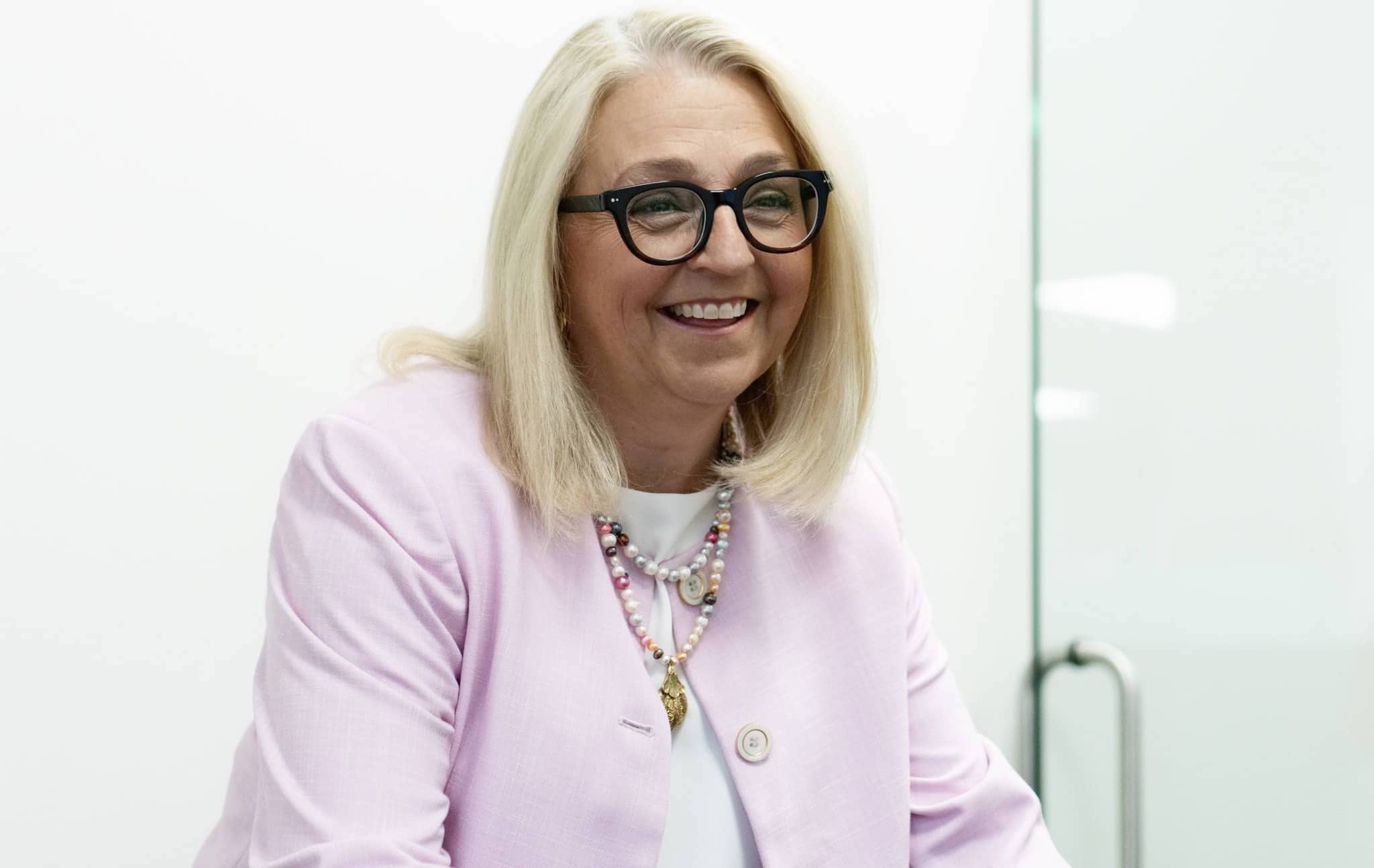 A smiling woman with long blonde hair, wearing black-framed glasses, a light pink blazer, and a beaded necklace. She is standing in front of a white wall with a partial view of a glass door in the background.