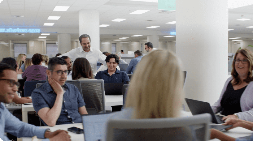 An office setting with multiple people working together at desks. Some people are seated and using laptops while others are standing and engaging in conversation. The environment is busy and collaborative, with a modern and professional atmosphere.