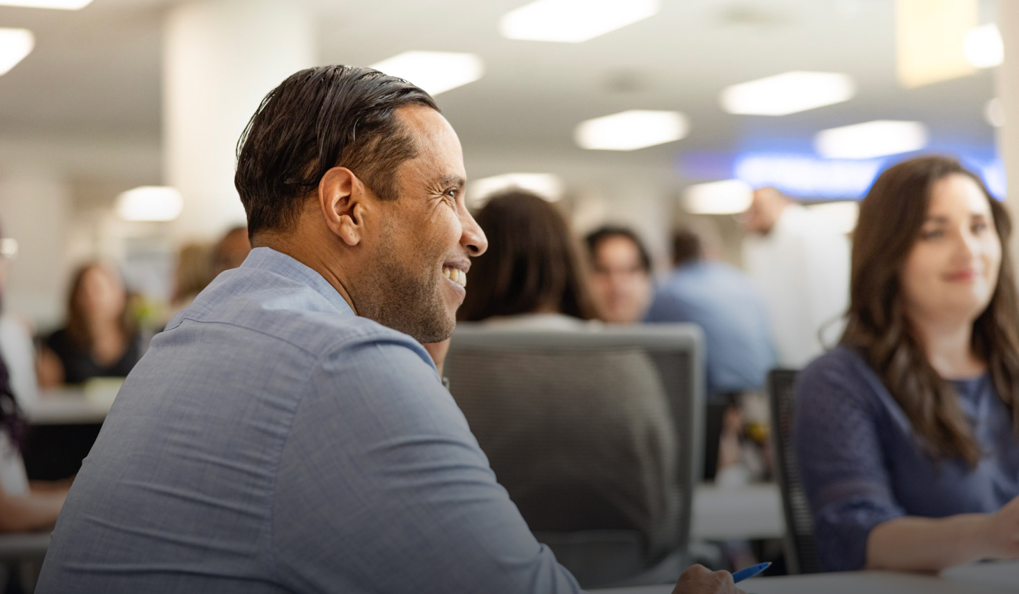 A man in a blue shirt smiles while sitting in an office environment with other people. The background shows a bright, busy workspace with soft lighting and individuals engaged in various activities.