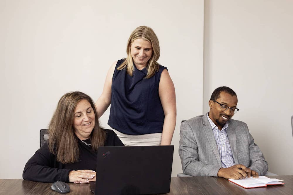 Three people are collaborating in an office setting. One woman sits at a desk working on a laptop, while another woman stands behind her, looking at the screen. A man is seated to the right, writing in a red notebook and smiling.